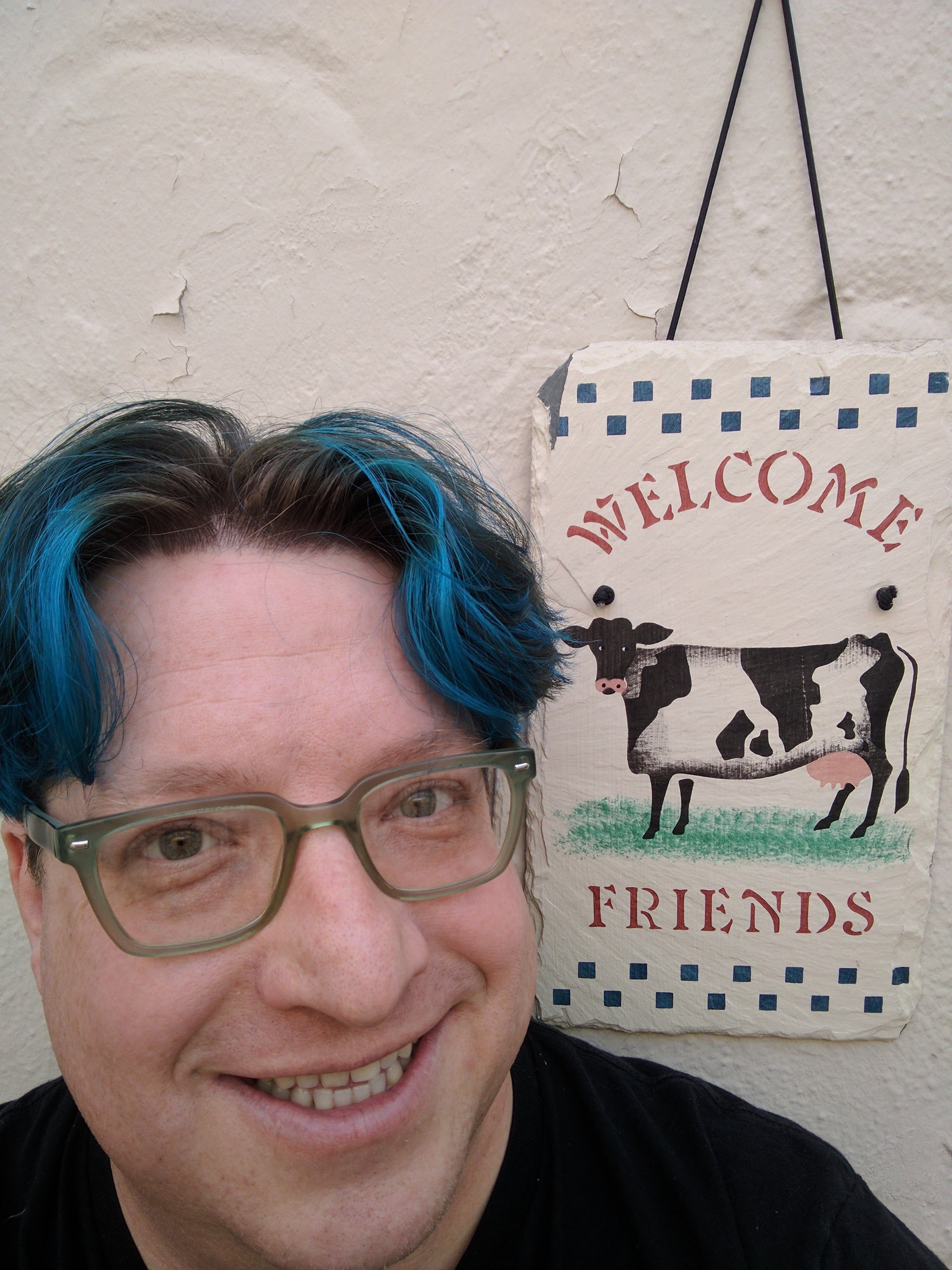 Blue-haired Eric next to a slate shingle with a cow and the word "welcome" painted upon it.